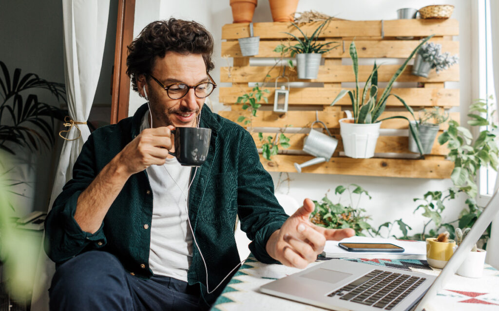 A man holding a cup, with plants in the background, doing hybrid remote work at his home office.