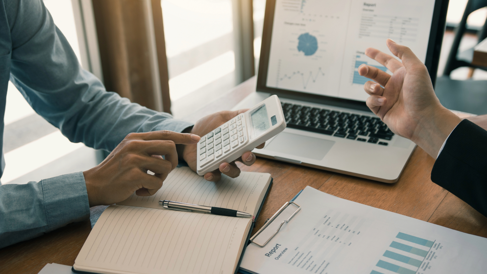 Two people discussing an IT budget at a desk with charts, a calculator, and a laptop displaying financial graphs.