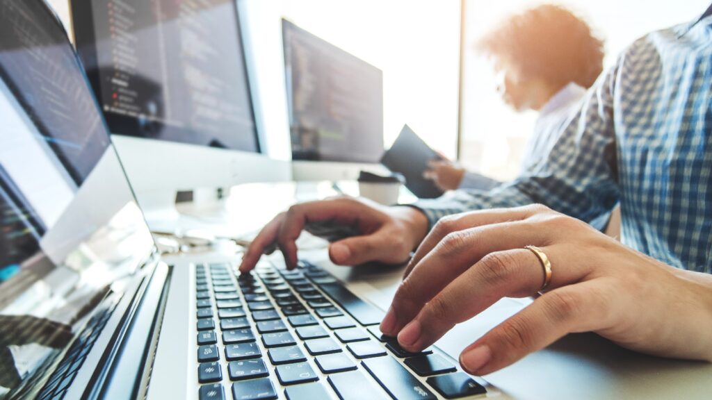 Close-up of a person typing on a laptop with computer monitors displaying code in the background, representing outsourced IT support.